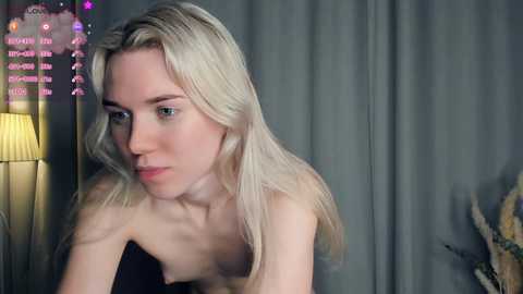 Video of a slender, pale-skinned blonde woman with wavy hair, wearing a strapless black top, looking contemplative in a dimly lit room with grey curtains and a lit lamp.