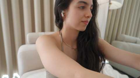 Video of a young woman with long black hair, fair skin, and a slender physique, wearing a white strapless dress, sitting on a beige couch. She has a neutral expression and is looking off to the side.