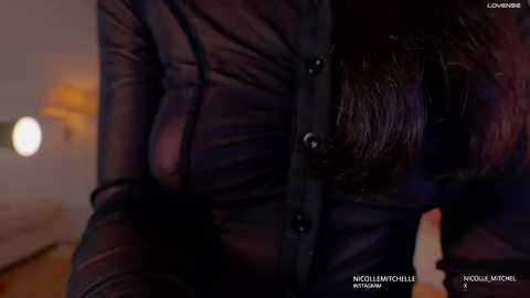 Video of a person wearing a sheer black leather jacket, highlighting the silhouette of a large, round breast beneath the fabric. Background features a dimly lit room with blurred, warm-toned furniture.