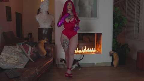 Video of a curvy woman with long red hair, wearing a red latex bodysuit and purple gloves, standing in a dimly-lit living room with a lit fireplace and decorative items.