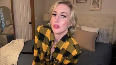 Video of a blonde woman with fair skin, wearing a yellow-green plaid shirt, sitting on a bed in a cozy bedroom with beige walls and framed pictures.