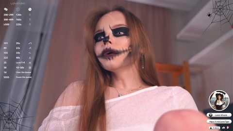 Video of a young woman with long brown hair, wearing a white off-shoulder top and heavy black makeup, including a black spiderweb across her face, in a dimly lit room with blurred furniture.