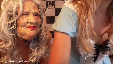 Video of an elderly woman with white, curly hair and a painted face resembling Alice in Wonderland, alongside a young girl in a blue dress with a black bow. Background has a black-and-white checkered pattern.