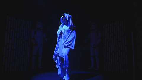 A video of a person dressed in a white robe with a hood, illuminated by blue light, standing in a dark, dimly lit room with vertical bars in the background.