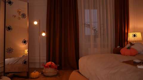 Video of a cozy, dimly-lit bedroom with a bed covered in cream sheets, a tall floor lamp, and a small white table with a pumpkin and flowers.