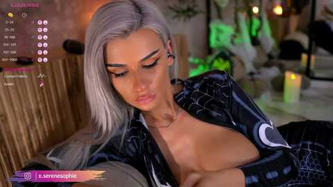 Video of a slender, platinum-blonde woman with high cheekbones, wearing a black latex dress, lying on a couch in a dimly lit, modern living room.