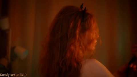 Video of a young woman with long, curly red hair and a black headband, viewed from the side in warm, dim lighting, showcasing her profile.