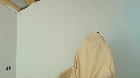 Video of a person with a beige robe, bending over, painting a plain white wall with exposed wooden beams in the background.