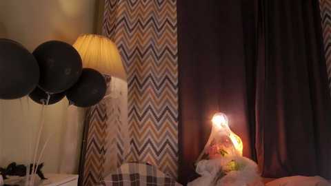 Video of a cozy bedroom corner with a black lampshade, black curtains, and a chevron-patterned curtain. A lit candle on a white table casts a warm glow, adding a romantic ambiance.