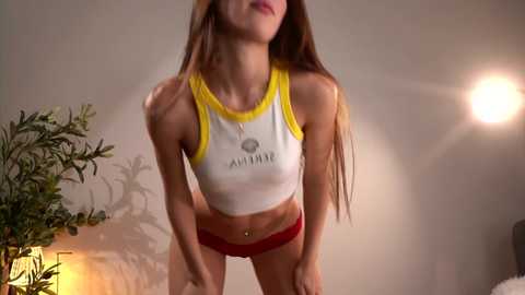 Video of a slender, fair-skinned woman with long brown hair, wearing a tight white crop top with yellow trim and red shorts, leaning forward in a dimly lit room.