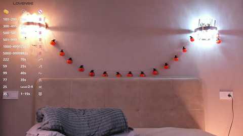 Video of a cozy bedroom with a beige tufted headboard, a gray and white striped blanket, and two wall-mounted, lit, pumpkin-shaped lamps on a cream-colored wall.