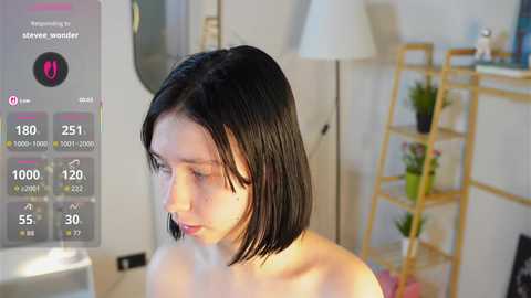 Video of a pale-skinned woman with shoulder-length black hair, topless, in a modern, minimalist room with a wooden shelf, a lamp, and a digital thermometer screen overlay.