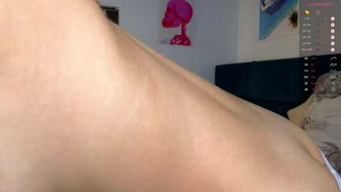 Video of a nude, light-skinned woman lying on her stomach, showcasing her back and shoulder blades. Background features a dark couch, pink lamp, and colorful artwork.