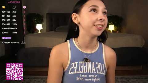 Video of a young woman with light skin and dark hair, wearing a blue sports tank top, smiling, in a dimly lit bedroom.