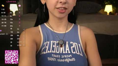 Video of a young Asian woman with long black hair, wearing a blue LA Lakers basketball jersey, a nose ring, and a pendant necklace. Background shows a dimly-lit bedroom with a bed and nightstand.