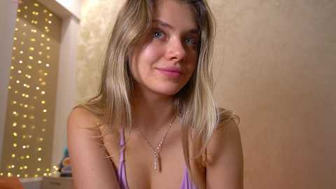 Video of a smiling, light-skinned woman with long, straight blonde hair, wearing a purple bralette and a delicate necklace, set against a warm, softly lit background with fairy lights.