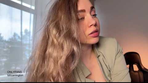 Video of a young woman with long, wavy, light brown hair, wearing a light green blazer, indoors by a window with natural light, looking contemplative. Text overlay reads \"ONLYFANS: talcompton.\