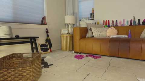 Video of a messy, modern living room with a beige leather couch, colorful vibrators, wicker baskets, and scattered clothes on a white tiled floor.