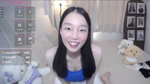 Media: Video of an Asian woman with long black hair, wearing a blue strapless top, sitting on a bed with plush toys.