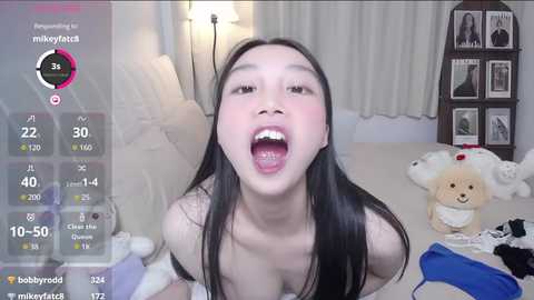 A video of a young Asian woman with long black hair, smiling widely, with a camera focused on her face, surrounded by a bedroom with a stuffed toy dog, clothes, and a bookshelf.