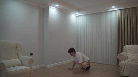 A video depicts a young man with short dark hair, wearing a white t-shirt and shorts, crouching on a light wooden floor in a minimalist, softly lit room with white walls, beige armchairs, and sheer white curtains.