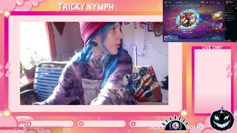 Video of a pale-skinned woman with blue hair, wearing a red beanie, purple striped sweater, and a colorful blanket, sitting in a cluttered room.