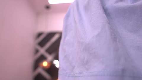 A video focuses on the shoulder and upper arm of a person wearing a light blue, slightly wrinkled shirt. The background is blurred, showing a dimly lit, geometric-patterned wall with soft lighting. The image has a soft focus and a warm color tone.