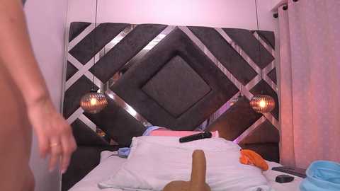 Video of a dimly lit bedroom with a modern, dark diamond-patterned headboard. A man and a woman, partially clothed, are in the foreground. The woman is lying on a bed, her legs visible, with a realistic, life-sized sex toy between them.