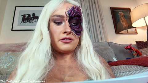 A video of a woman with long, platinum blonde hair and a purple, eye-like prosthetic covering her right eye, sitting on a gray couch in a living room with framed art and a lamp.
