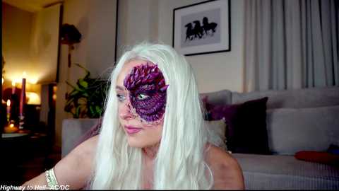 Video of a fair-skinned woman with long platinum blonde hair, wearing a purple, feathered masquerade mask, sitting on a beige couch in a dimly-lit living room.