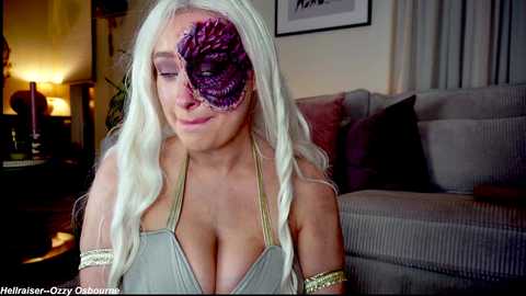 A video shows a blonde woman with a large, purple, eye-like prosthetic on her forehead, wearing a low-cut, gray top. She sits in a dimly lit living room with a gray sofa and a lamp.
