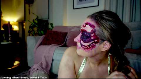 Video of a woman with a Halloween-themed face paint, featuring a white skull with red and black details, wearing a gold top, in a dimly lit living room with a lamp, cushions, and framed art on the wall.