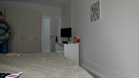 Media: Video of a minimalist, well-lit bedroom with beige walls and a white desk against the right wall, featuring a computer monitor and a colorful abstract painting. The bed, with a beige comforter, occupies the foreground.