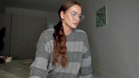 Media: Video of a young woman with long brown hair, wearing glasses and a gray-striped sweater, standing indoors next to a white wall with a green abstract painting.