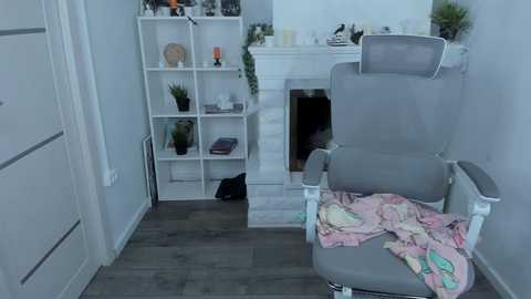 A video of a modern, minimalist living room with a gray massage chair, white shelves, a white fireplace, and a white door. The room has a clean, neutral color palette with soft lighting.