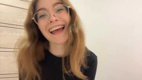 Video of a Caucasian woman with long, wavy blonde hair, wearing round glasses, and a black top, smiling widely with her mouth open, against a beige wooden wall.