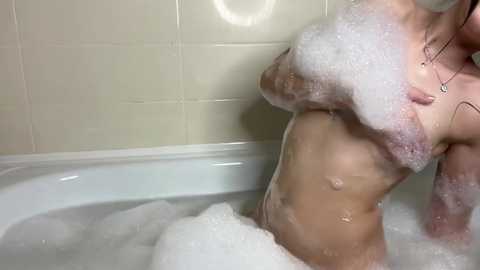 Media: Video of a partially submerged, naked woman with fair skin and wet hair, using her left hand to cover her right breast, bubbles covering her body in a white bathtub.