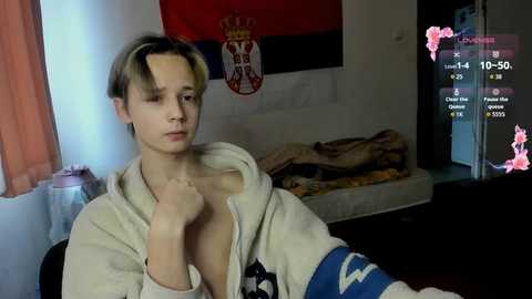 Video of a young Asian man with short, blonde hair, wearing a hoodie, in a dimly lit bedroom with a bed, a flag, and a pink overlay on the right side.