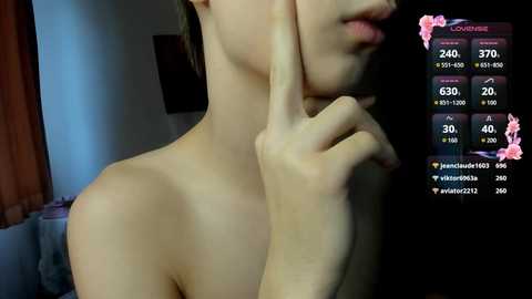 A close-up video of a topless woman with light skin, using her finger to cover her mouth, with a smartphone screen showing live chat details in the background.