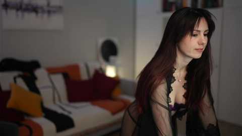 Video of a pale, slender woman with long dark hair, wearing a sheer black robe, standing in a modern living room with a couch, pillows, and abstract art.