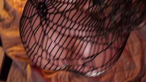Video of a close-up view of a person wearing a black fishnet top, revealing their large, round breasts and a tattoo on their right breast. Background features a warm, textured fabric.
