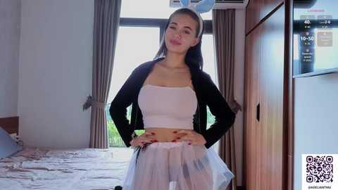 Media: Video of a young Asian woman with long black hair, wearing a white crop top, sheer skirt, and black cardigan, standing in a modern, brightly lit room with wooden furniture and a bed.