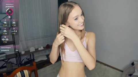 Media: Video of a slim, light-skinned young woman with long blonde hair, wearing a light pink bra, smiling while adjusting her hair in a bedroom with a bed, window, and TV.