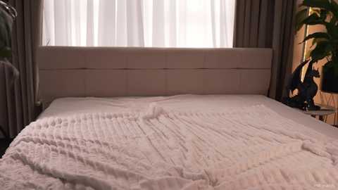 Video of a modern bedroom with a plush, white, quilted bedspread, beige upholstered headboard, sheer white curtains, and a dark wooden nightstand with a black statue.