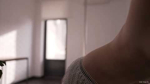 Video of a close-up of a woman's bare shoulder and upper arm, wearing a shimmering, off-shoulder top. The background is a softly lit hallway with white walls and a glass door.