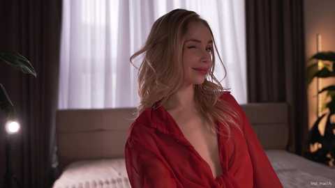 Video of a smiling, fair-skinned blonde woman with wavy hair, wearing a red sheer blouse, standing in a dimly lit bedroom with white curtains and a bed in the background.