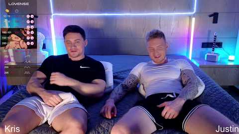 Video of two muscular men in tight shirts and shorts, lying on a bed with a gray blanket. Background features a sleek, modern bedroom with blue LED lighting.