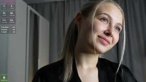 Video of a young, fair-skinned woman with blonde hair pulled back, wearing a black top, smiling softly. Background features gray curtains and a partially visible door.