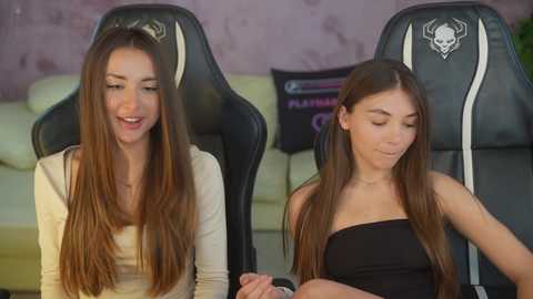 Video of two young women with long, straight brown hair, seated on gaming chairs in a room with a light purple wall and a white couch. One wears a white top, the other a black strapless dress.