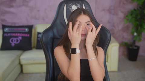 Video of a young woman with long brown hair, wearing a black top and a smartwatch, sitting on a gaming chair in a room with a cream couch, purple wall, and green potted plant.